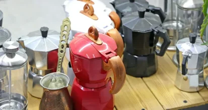 A shelf holding several different manual coffee brewers side by side: colorful stovetop moka pots, a glass French press, a hand grinder and a small copper cezve