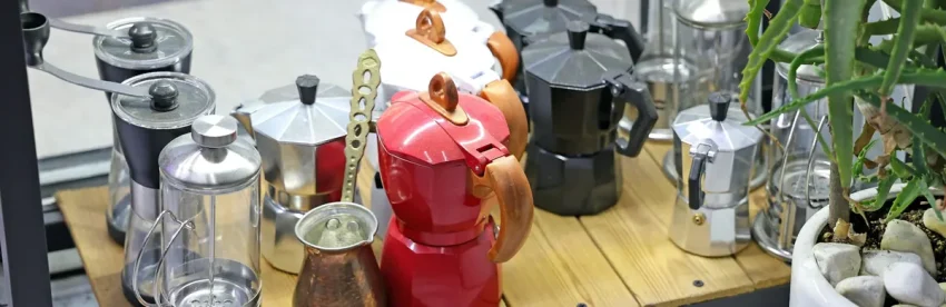 A shelf holding several different manual coffee brewers side by side: colorful stovetop moka pots, a glass French press, a hand grinder and a small copper cezve