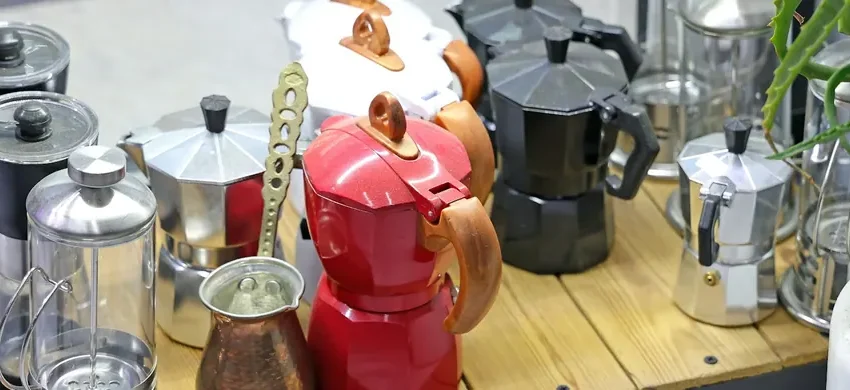 A shelf holding several different manual coffee brewers side by side: colorful stovetop moka pots, a glass French press, a hand grinder and a small copper cezve