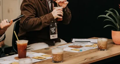 Two competition judges closely evaluating and tasting drinks, with scoring cards visible on the table in front of them