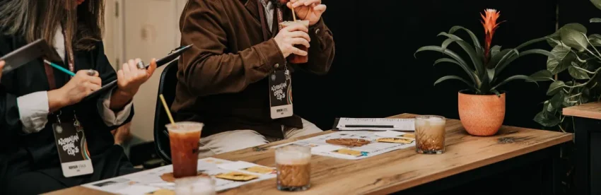 Two competition judges closely evaluating and tasting drinks, with scoring cards visible on the table in front of them