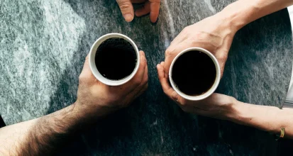 Overhead view of two hands holding cups of black coffee over a stone table