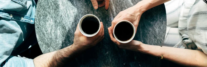 Overhead view of two hands holding cups of black coffee over a stone table