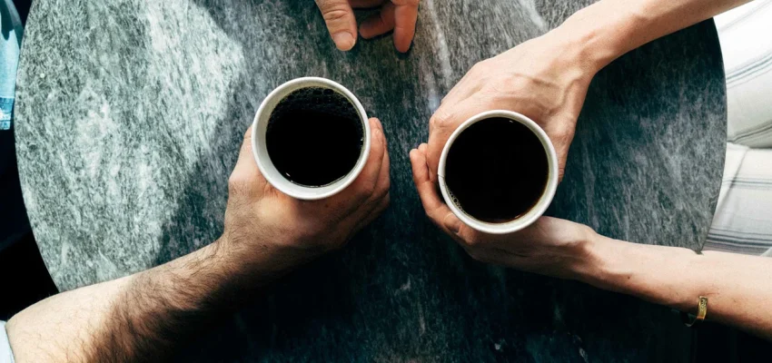 Overhead view of two hands holding cups of black coffee over a stone table