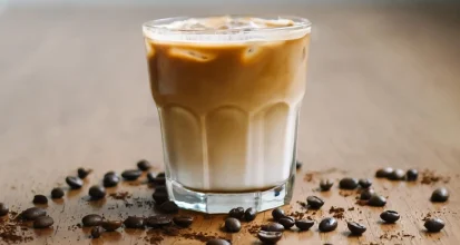 Iced Brazilian latte in a short glass tumbler showing a pale condensed milk layer at the bottom and caramel coloured coffee above, with roasted beans and ground coffee scattered on a wooden table
