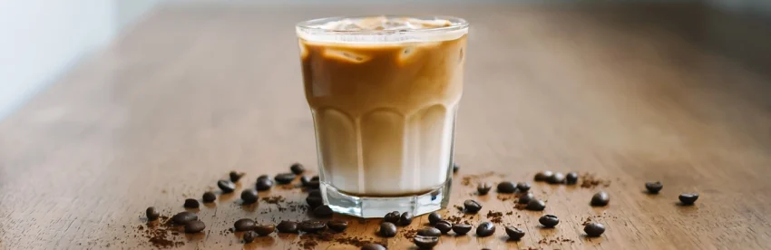 Iced Brazilian latte in a short glass tumbler showing a pale condensed milk layer at the bottom and caramel coloured coffee above, with roasted beans and ground coffee scattered on a wooden table