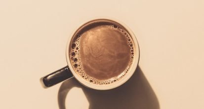 Overhead view of a single dark mug of black coffee on a beige background, casting a long shadow to the right