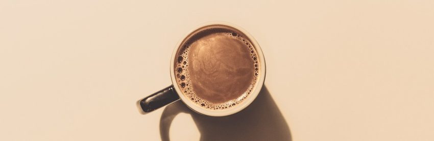 Overhead view of a single dark mug of black coffee on a beige background, casting a long shadow to the right