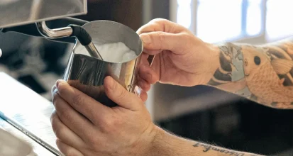 A barista with a tattooed arm steaming milk in a stainless steel pitcher at an espresso machine steam wand
