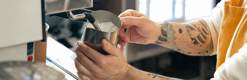 A barista with a tattooed arm steaming milk in a stainless steel pitcher at an espresso machine steam wand