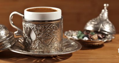 A cup of foamy Turkish coffee in an ornate engraved silver cup holder on a matching saucer, with a lidded silver sugar dish and a small bowl of colorful Turkish delight on a wooden table