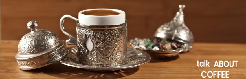 A cup of foamy Turkish coffee in an ornate engraved silver cup holder on a matching saucer, with a lidded silver sugar dish and a small bowl of colorful Turkish delight on a wooden table