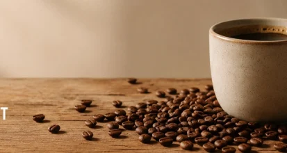 A steaming ceramic mug of black coffee on a rustic wooden table with roasted coffee beans scattered in front, against a warm neutral background