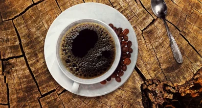 Overhead view of a cup of black coffee on a rustic wooden surface with scattered coffee beans and a spoon