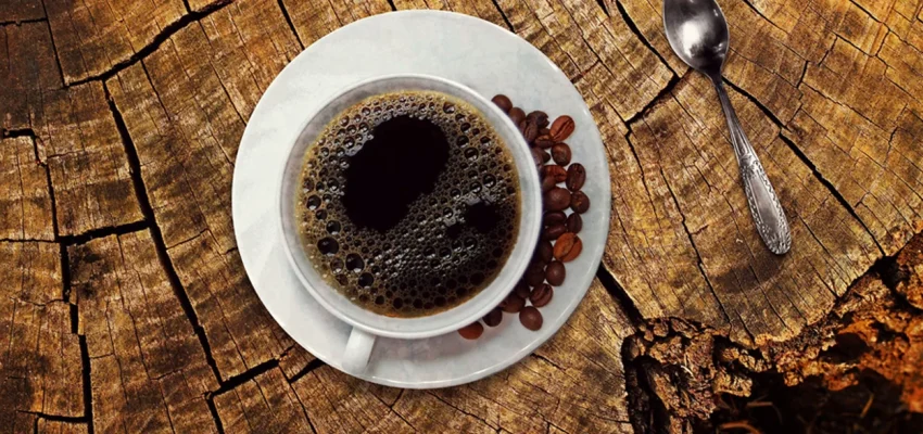 Overhead view of a cup of black coffee on a rustic wooden surface with scattered coffee beans and a spoon