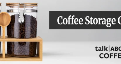 Whole coffee beans being poured from a kraft bag into an opaque airtight storage canister on a wooden kitchen counter