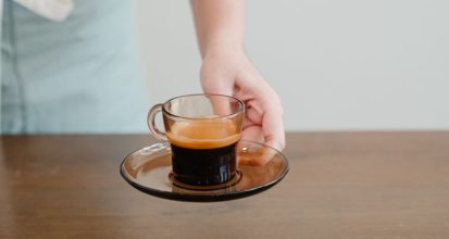 Barista in an apron setting down a glass cup of espresso with crema on a saucer, on a wooden counter beside a tamper