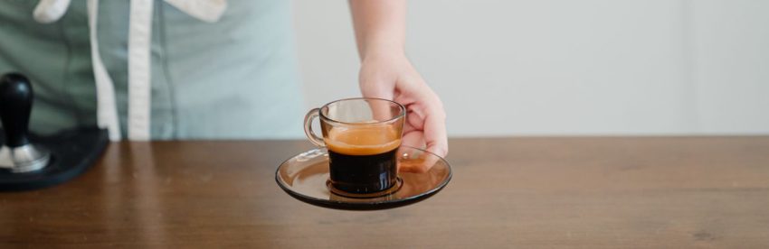 Barista in an apron setting down a glass cup of espresso with crema on a saucer, on a wooden counter beside a tamper