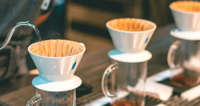 Row of identical white pour-over drippers with paper filters on glass cups, water being poured from a gooseneck kettle into the first one