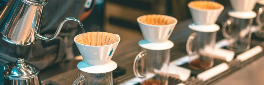 Row of identical white pour-over drippers with paper filters on glass cups, water being poured from a gooseneck kettle into the first one