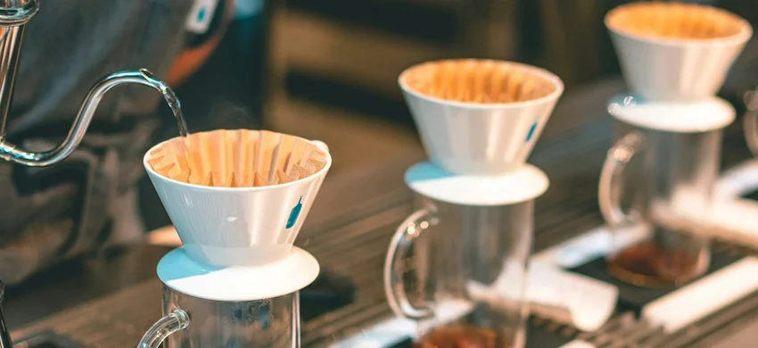 Row of identical white pour-over drippers with paper filters on glass cups, water being poured from a gooseneck kettle into the first one