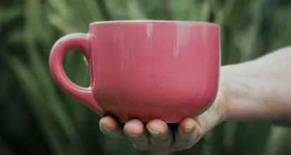 A hand holding a pink mug of coffee against a background of green leaves