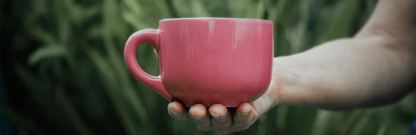A hand holding a pink mug of coffee against a background of green leaves
