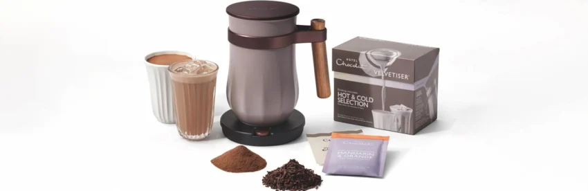 Electric hot chocolate maker beside a hot and an iced chocolate drink, a box of drinking chocolate sachets, cocoa powder and chocolate flakes