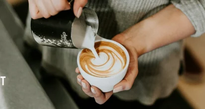 Overhead view of a barista pouring steamed milk from a metal pitcher into a white cup to create a rosetta latte-art pattern