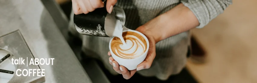 Overhead view of a barista pouring steamed milk from a metal pitcher into a white cup to create a rosetta latte-art pattern
