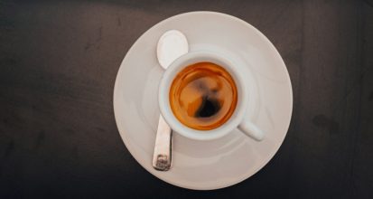 Overhead view of a shot of espresso with golden crema in a white cup and saucer with a spoon, on a dark charcoal background