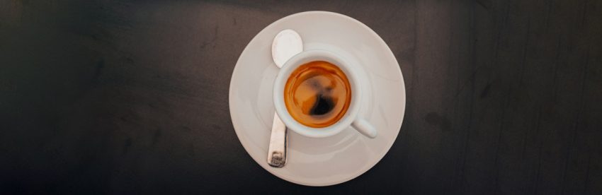 Overhead view of a shot of espresso with golden crema in a white cup and saucer with a spoon, on a dark charcoal background
