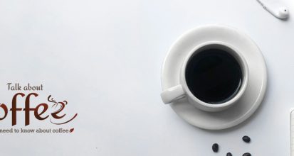 Overhead view of a white cup of black coffee on a saucer beside a spiral notepad, pen, scattered coffee beans and earphones on a pale grey desk