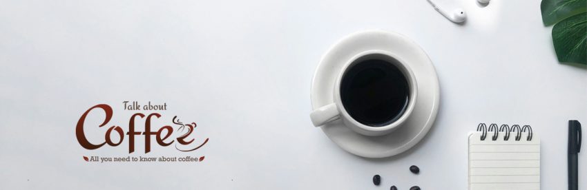 Overhead view of a white cup of black coffee on a saucer beside a spiral notepad, pen, scattered coffee beans and earphones on a pale grey desk