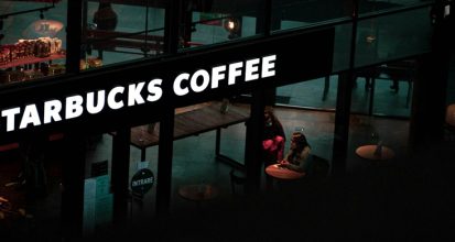 Glowing Starbucks Coffee sign over a dimly lit store interior with customers seated at tables, viewed from above at night