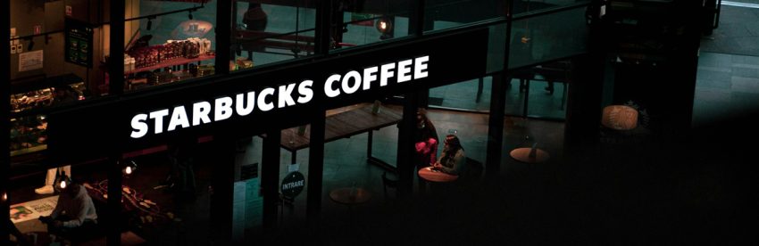 Glowing Starbucks Coffee sign over a dimly lit store interior with customers seated at tables, viewed from above at night