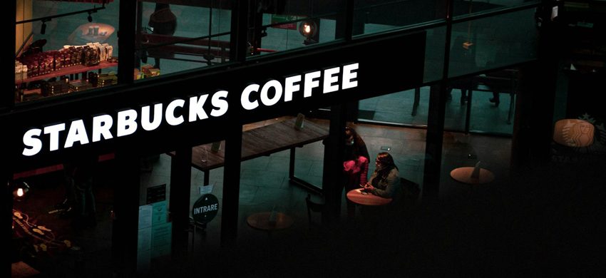 Glowing Starbucks Coffee sign over a dimly lit store interior with customers seated at tables, viewed from above at night