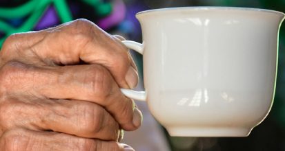 The weathered hand of an older woman holding a white china coffee cup by the handle, against a colourful blurred background