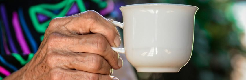 The weathered hand of an older woman holding a white china coffee cup by the handle, against a colourful blurred background