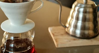 A ceramic pour-over dripper sitting on a glass carafe of freshly brewed coffee, next to a gooseneck kettle on a kitchen table