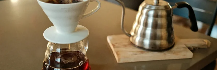 A ceramic pour-over dripper sitting on a glass carafe of freshly brewed coffee, next to a gooseneck kettle on a kitchen table