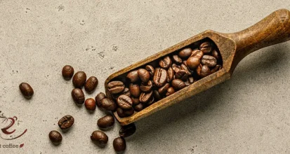 Dark roasted coffee beans spilling from a wooden scoop onto a pale stone surface, shot from above