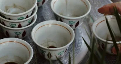 A tray of small ceremonial coffee cups with dark coffee poured into several of them, a hand reaching in among tall grass stems