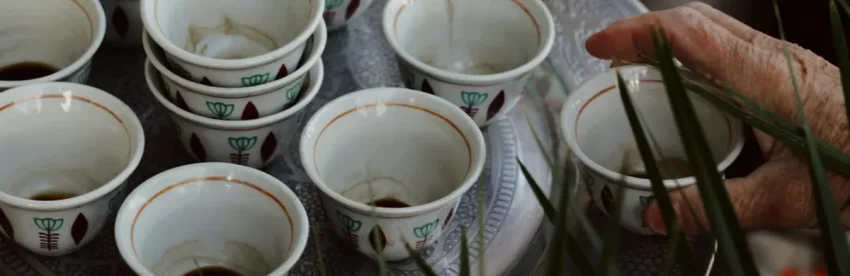 A tray of small ceremonial coffee cups with dark coffee poured into several of them, a hand reaching in among tall grass stems