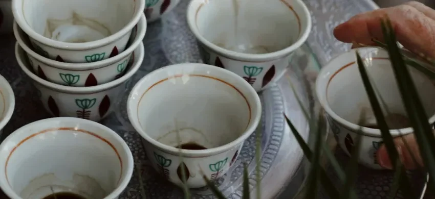 A tray of small ceremonial coffee cups with dark coffee poured into several of them, a hand reaching in among tall grass stems