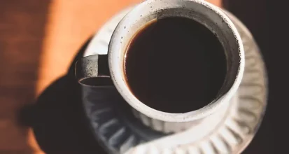 An overhead view of a white cup filled with black coffee on a wooden table, lit by a warm diagonal beam of morning sunlight