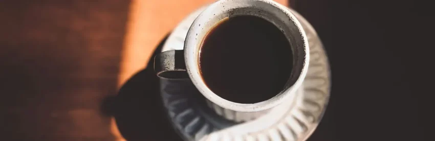 An overhead view of a white cup filled with black coffee on a wooden table, lit by a warm diagonal beam of morning sunlight