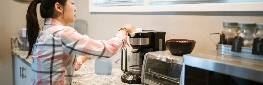 A person in a kitchen wiping down the outside of a drip coffee maker with a cloth as part of a regular cleaning routine
