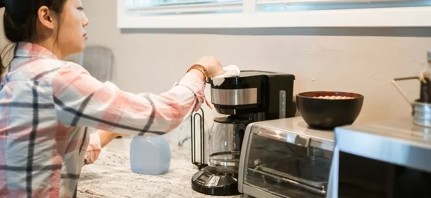 A person in a kitchen wiping down the outside of a drip coffee maker with a cloth as part of a regular cleaning routine