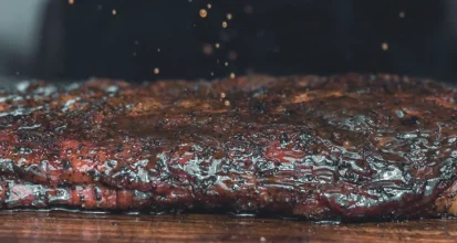 Close-up of a coffee-rubbed brisket with a dark glossy bark crust resting on a wooden cutting board, with spices falling through the air